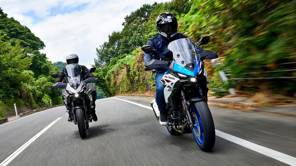 Zwei Motorradfahrer auf einer kurvigen Straße; links ein graues Motorrad, rechts ein blau-weißes Suzuki-Motorrad. Üppiges Grün auf beiden Straßenseiten.