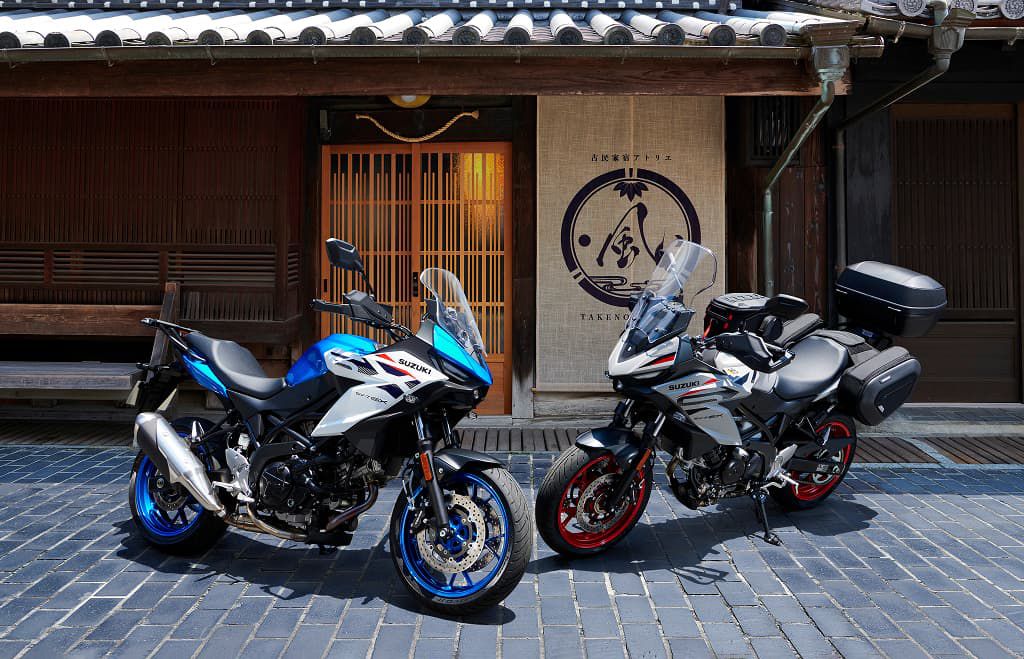 Zwei Suzuki-Motorräder stehen nebeneinander vor einem traditionellen japanischen Gebäude; links ein blau-weißes Modell, rechts ein grau-weißes Modell mit Touring-Koffern.