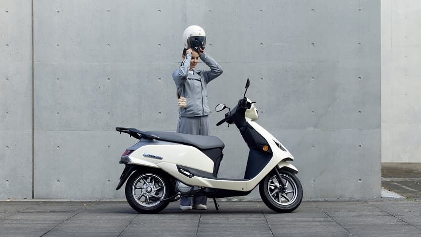 Eine Person setzt sich einen weißen Helm auf und steht neben einem weißen Suzuki e-Address Elektroroller vor einer grauen Betonwand.