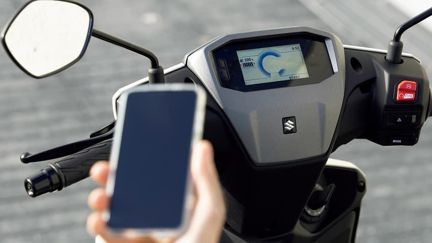 Nahaufnahme des Cockpits eines Suzuki e-Address; im Vordergrund hält jemand ein Smartphone, im Hintergrund ist das digitale Display des Rollers zu sehen.