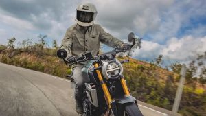 Motorradfahrer mit beiger Jacke und weißem Helm fährt eine Suzuki GSX-8T auf einer Landstraße bei bewölktem Himmel