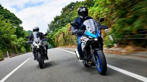 Zwei Motorradfahrer auf einer kurvigen Straße; links ein graues Motorrad, rechts ein blau-weißes Suzuki-Motorrad. Üppiges Grün auf beiden Straßenseiten.