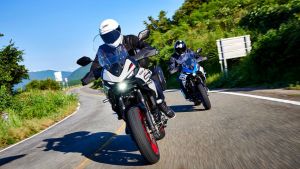 Zwei Motorradfahrer fahren zügig durch eine kurvige Landstraße; im Vordergrund ein weißes Adventure-Bike mit roten Felgen, dahinter ein blaues Motorrad. Grüne Vegetation und Berge im Hintergrund.