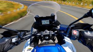 Perspektive aus der Fahreransicht eines Motorrads auf einer Landstraße; Hände am Lenker, digitales Display sichtbar, die Straße verläuft leicht kurvig nach vorne.