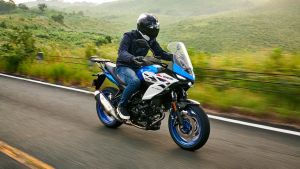 Ein Motorradfahrer fährt ein blau-weißes Suzuki-Motorrad auf einer Landstraße; grüne Hügel im Hintergrund und weiches Abendlicht.