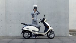 Eine Person setzt sich einen weißen Helm auf und steht neben einem weißen Suzuki e-Address Elektroroller vor einer grauen Betonwand.