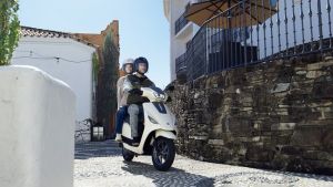 Zwei Personen fahren auf einem weißen Suzuki e-Address Elektroroller durch eine enge, gepflasterte Gasse, flankiert von weißen Fassaden und einer Natursteinmauer.