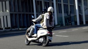 Zwei Personen fahren auf einem weißen Suzuki e-Address Elektroroller durch eine moderne Innenstadt mit hohen Glas- und Betonfassaden.