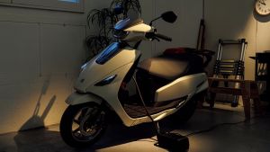 Ein weißer Suzuki e-Address Elektroroller steht in einer Garage und wird über ein Ladekabel geladen; gedämpftes Licht und Gegenstände im Hintergrund.