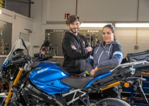 Ein Mechaniker und eine Mechanikerin stehen mit verschränkten Armen vor einem blauen Suzuki-Motorrad in der Werkstatt.