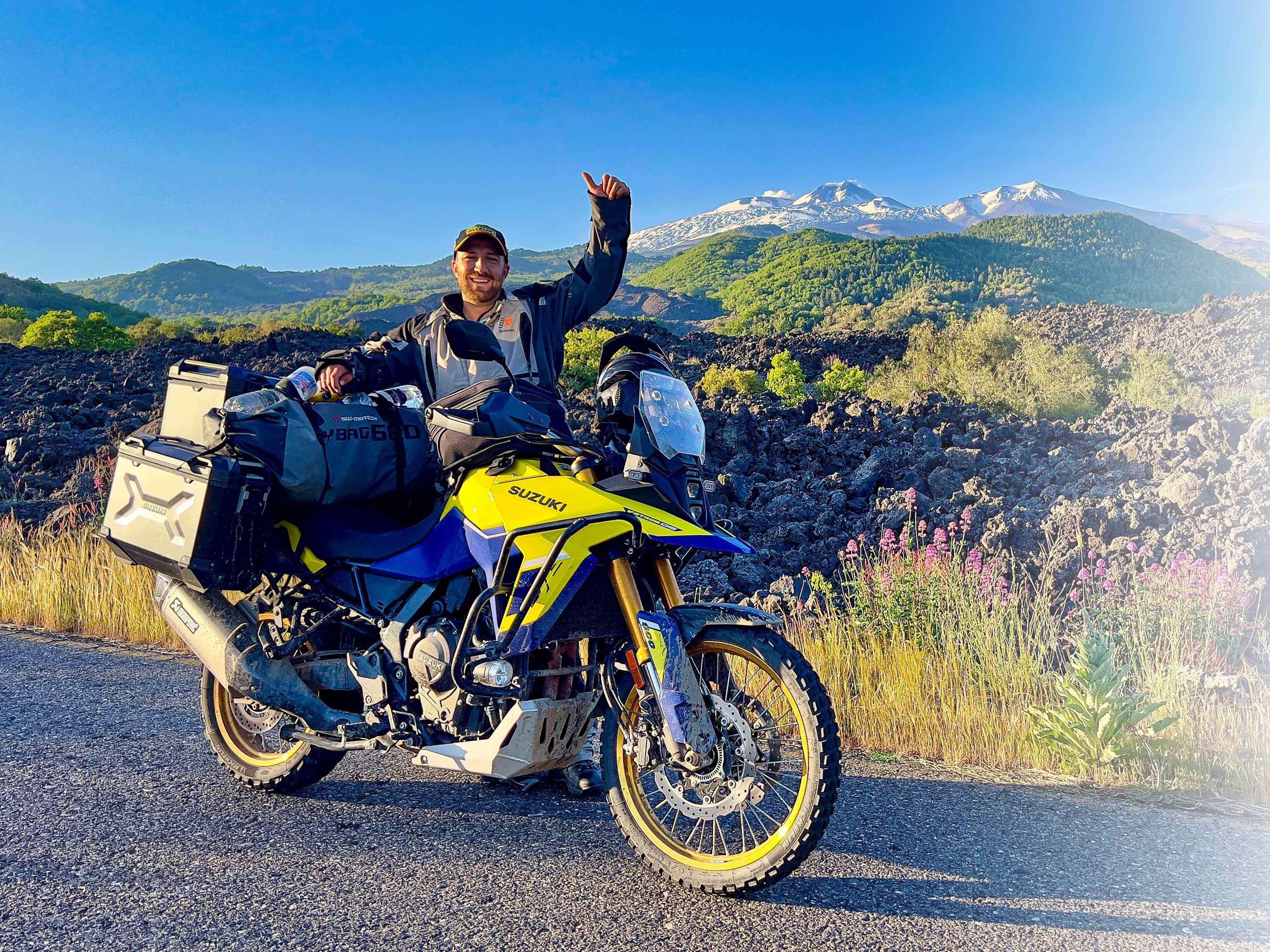 Offroad-Abenteurer Stefan Meitinger mit seiner Suzuki V-Strom 800DE, beladen mit Gepäck und Seitenkoffern, posiert auf einer Straße vor einer beeindruckenden Berglandschaft mit grünen Hügeln und schneebedeckten Gipfeln.