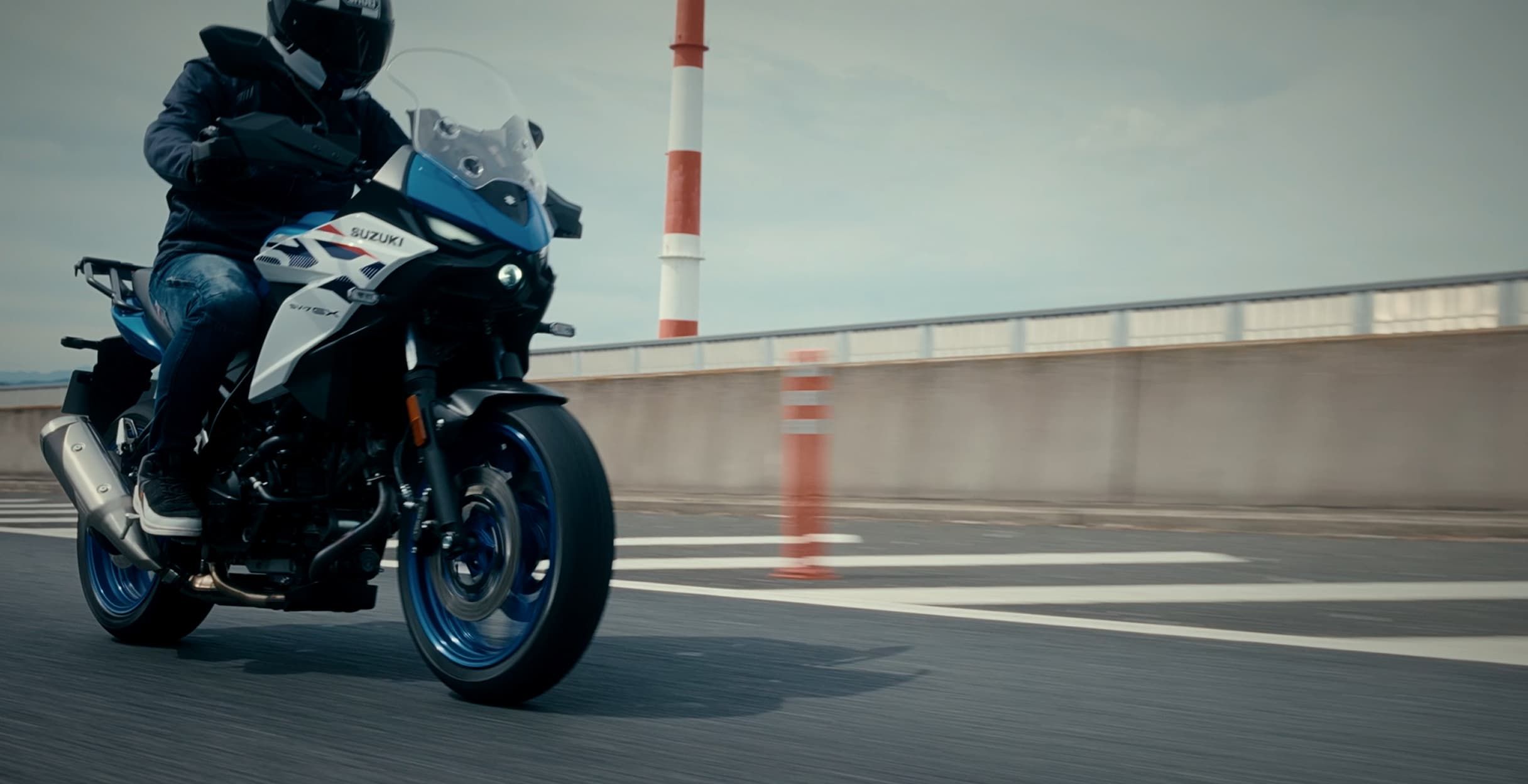 Ein Motorradfahrer fährt ein Suzuki-Motorrad auf einer Autobahn; Fokus auf das Vorderrad, die Verkleidung und die Bewegung des Motorrads, mit Leitplanke und trübem Himmel im Hintergrund.