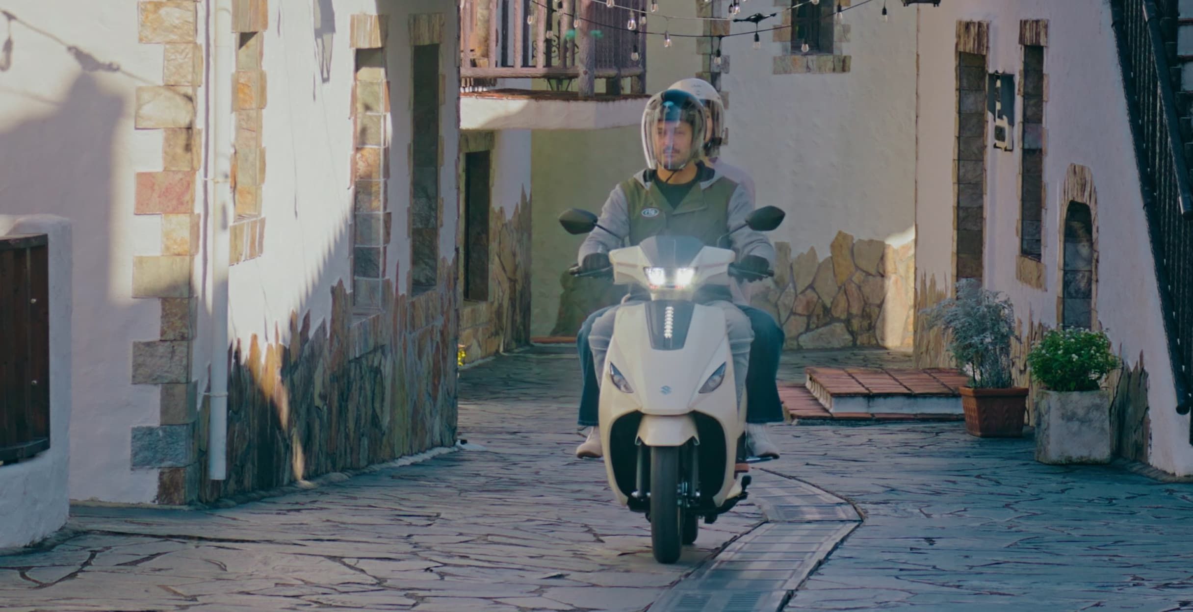 Zwei Personen fahren auf einem weißen Suzuki e-Address Elektroroller durch eine enge, gepflasterte Gasse mit weiß verputzten Häusern und Natursteinfassaden.