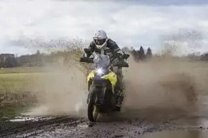 Motorradfahrer auf Suzuki V-Strom 800DE fährt durch schlammige Pfütze in ländlicher Umgebung.