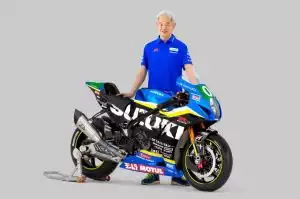 Mann in blauem Hemd steht lächelnd neben einer blauen, schwarz-gelben Suzuki Rennmaschine mit Sponsorenlogos vor grauem Hintergrund.