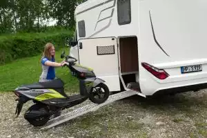 Frau lädt Suzuki Avenis Roller in Wohnmobil, grüne Wiese im Hintergrund