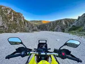 Motorradfahrt durch felsige Landschaft bei Sonnenuntergang, Fokus auf Lenker und Display.