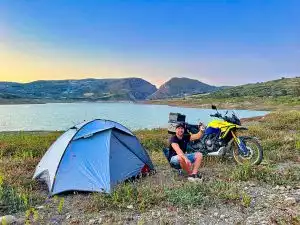 Mann neben Suzuki-Motorrad und Zelt am Seeufer in bergiger Landschaft