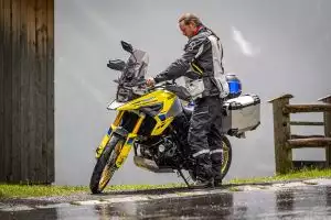 Motorradfahrer mit Suzuki V-Strom 1050XT auf nasser Straße vor Holzhütte