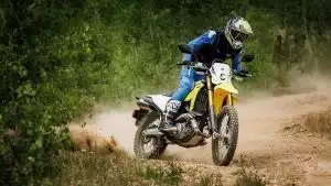 Motorradfahrer auf gelber Suzuki DR-Z4S im Gelände, umgeben von Bäumen.