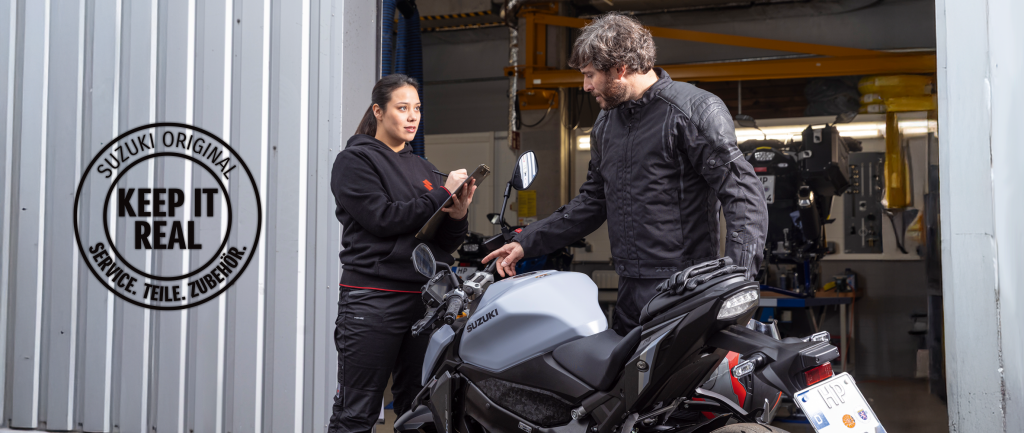 Suzuki Service-Mitarbeiterin im Gespräch mit einem Kunden an seiner grauen Suzuki GSX-S950 vor der Werkstatt. Links im Bild das runde Logo „Suzuki Original – Keep it real. Service. Teile. Zubehör.“ als Hinweis auf originalgetreuen Service und Ersatzteile.
