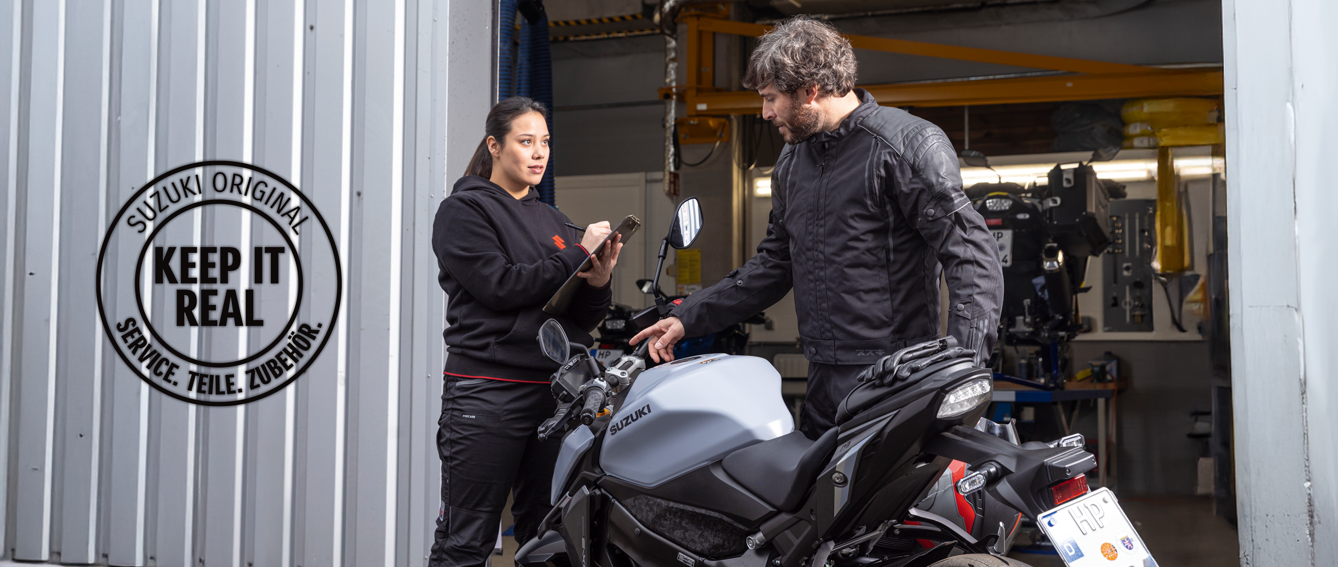 Suzuki Service-Mitarbeiterin im Gespräch mit einem Kunden an seiner grauen Suzuki GSX-S950 vor der Werkstatt. Links im Bild das runde Logo „Suzuki Original – Keep it real. Service. Teile. Zubehör.“ als Hinweis auf originalgetreuen Service und Ersatzteile.