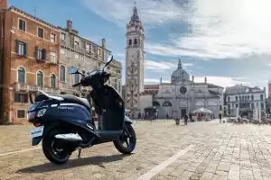 Der Suzuki Address 125 in mattblauer Lackierung steht auf einem gepflasterten Platz vor der barocken Fassade einer historischen Kirche mit Uhrturm.