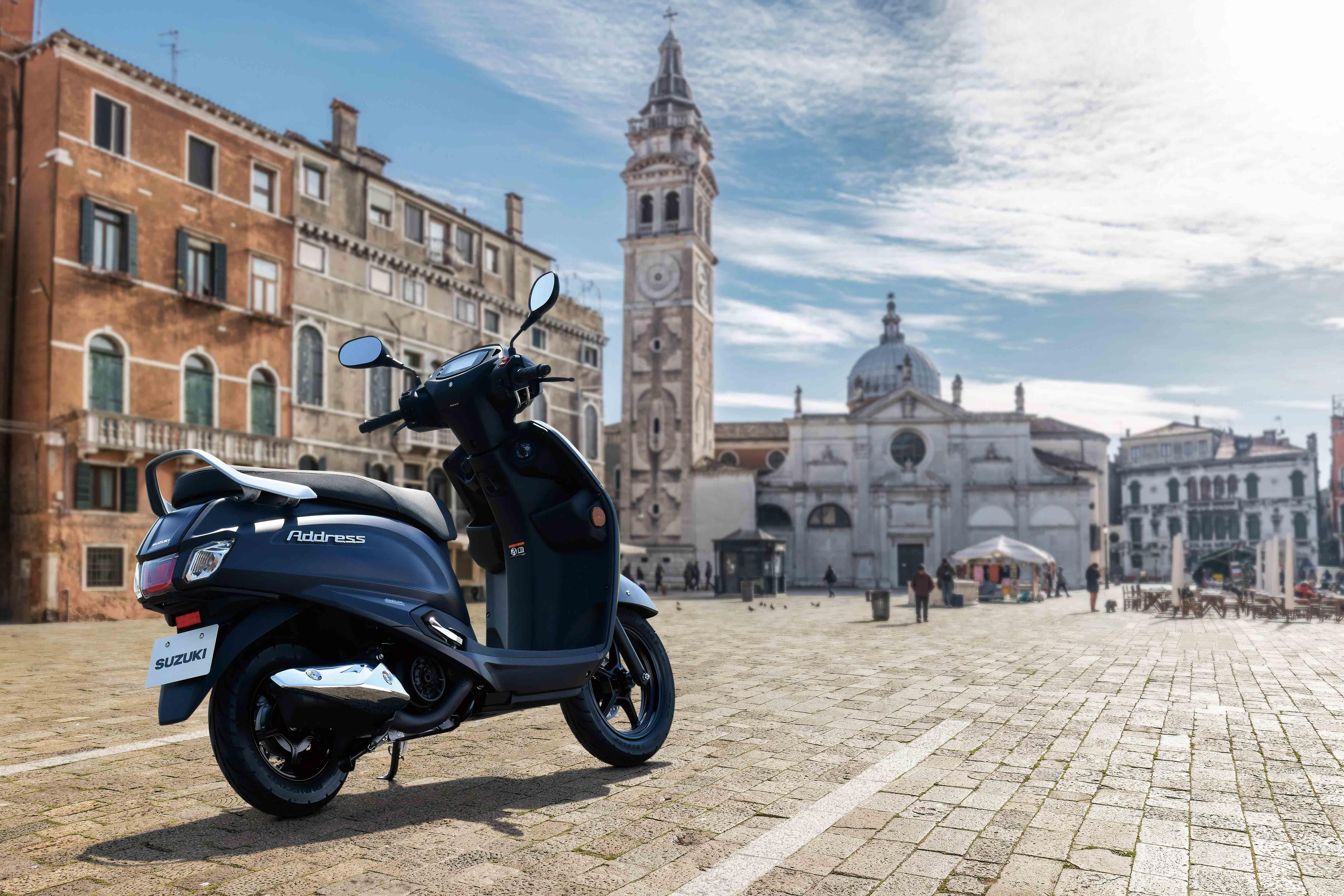 Der Suzuki Address 125 in mattblauer Lackierung steht auf einem gepflasterten Platz vor der barocken Fassade einer historischen Kirche mit Uhrturm.