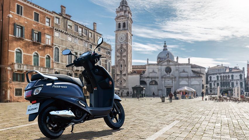 Der Suzuki Address 125 in mattblauer Lackierung steht auf einem gepflasterten Platz vor der barocken Fassade einer historischen Kirche mit Uhrturm.