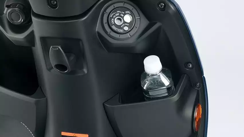 Nahaufnahme des vorderen Innenraums des Suzuki Address 125 mit Blick auf das Zündschloss, eine offene Ablage mit eingesetzter Wasserflasche, einer 12V-Steckdose und Taschenhaken.