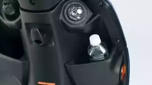 Nahaufnahme des vorderen Innenraums des Suzuki Address 125 mit Blick auf das Zündschloss, eine offene Ablage mit eingesetzter Wasserflasche, einer 12V-Steckdose und Taschenhaken.