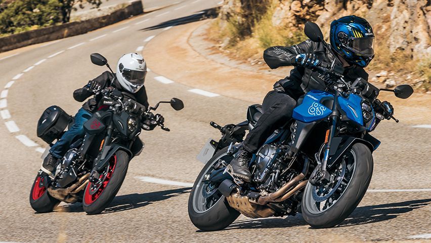 Zwei Suzuki GSX-8S in sportlicher Schräglage auf einer kurvigen Landstraße, vorne in Blau, dahinter ein schwarzes Modell mit roten Felgen.