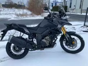 Seitenansicht von einem Suzuki Motorrad im Schnee mit Stollenreifen, Windschutzscheibe und goldener Upside-down-Gabel.