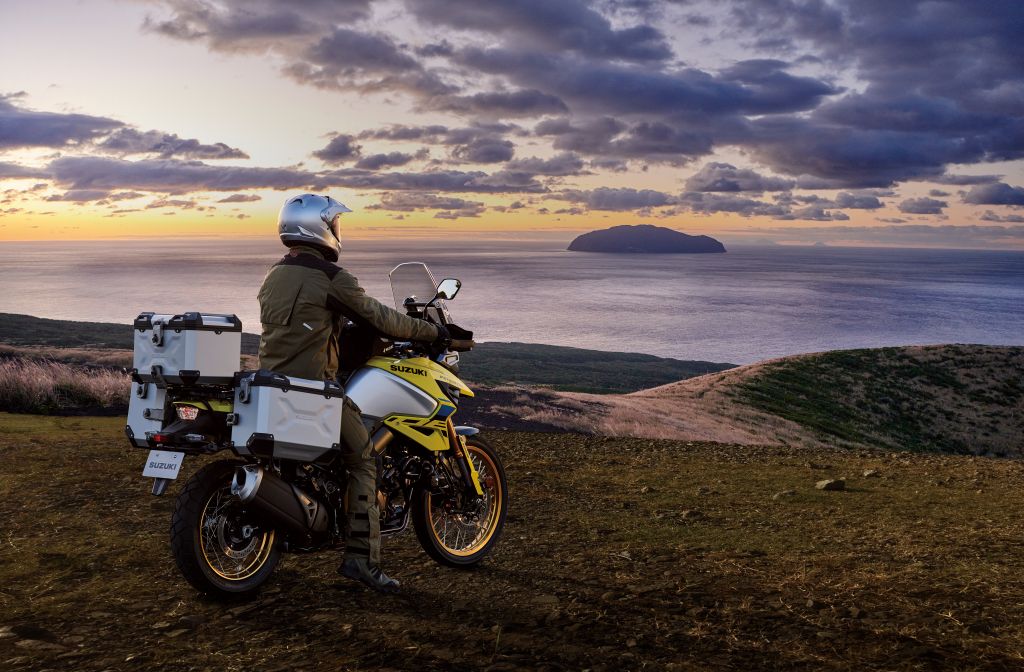 Mann mit silbernem Helm und dunkelgrüner Jacke sitzt auf einem gelben Suzuki V-Strom 1050DE Modell von 2024 mit Aluminiumkoffer. Das Motorrad steht auf einer erhöhten Wiese von wo aus der Mann das Meer und den Sonnenaufgang im Hintergrund betrachtet.