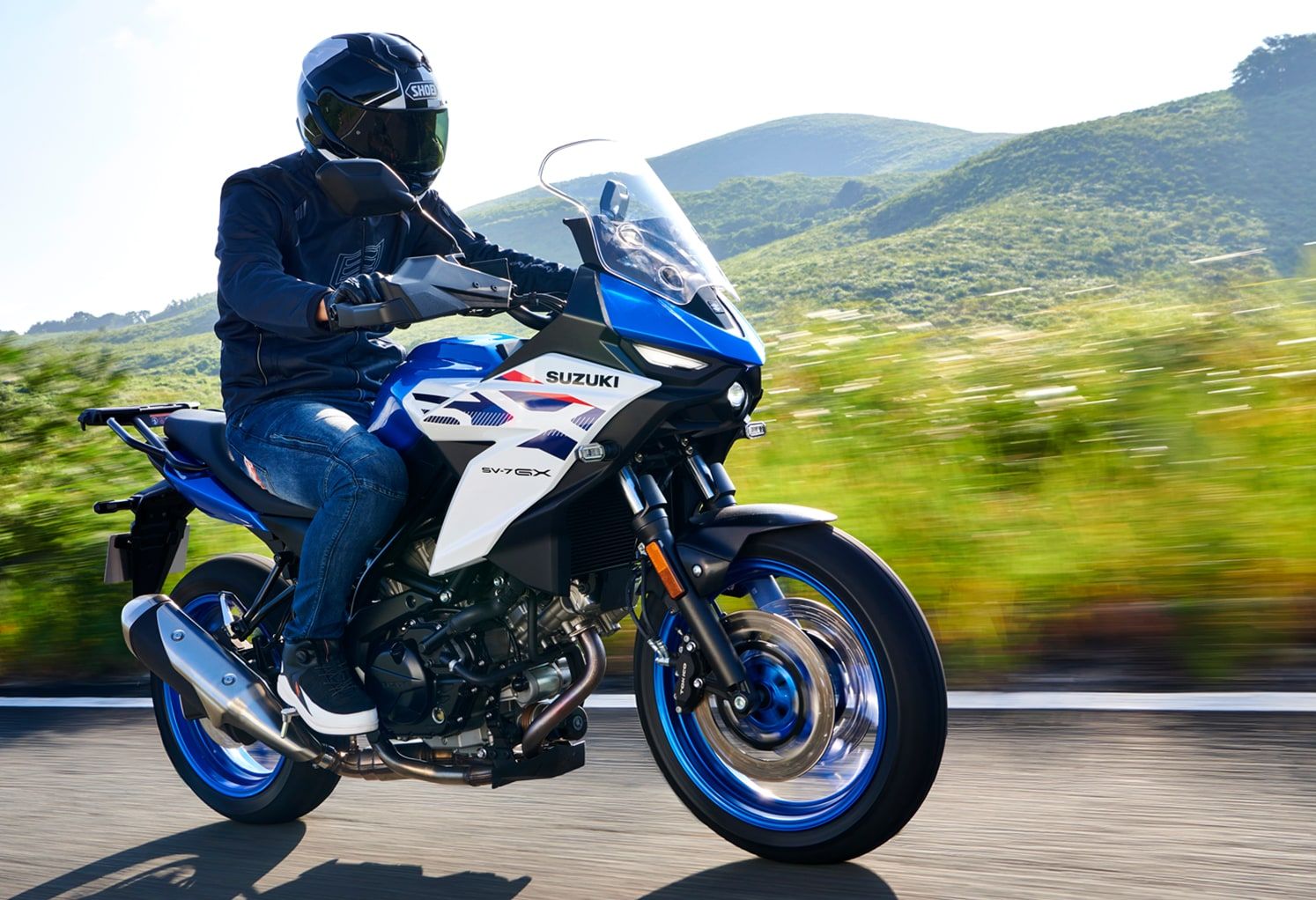 Eine Person mit schwarzem Motorradhelm fährt auf einer blau-weißen Suzuki SV-7GX über eine Landstraße vor grünen Hügeln