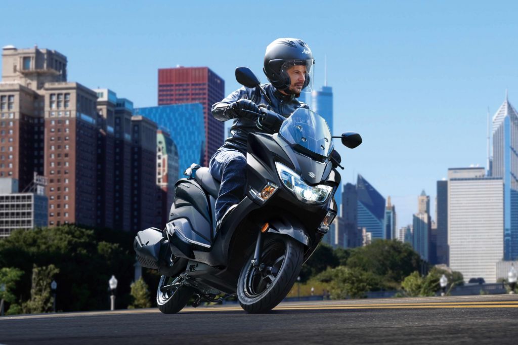 Fahrer mit auf einem Suzuki BURGMAN STREET 125EX unterwegs durch eine moderne Großstadt, umgeben von Hochhäusern und klarblauem Himmel.