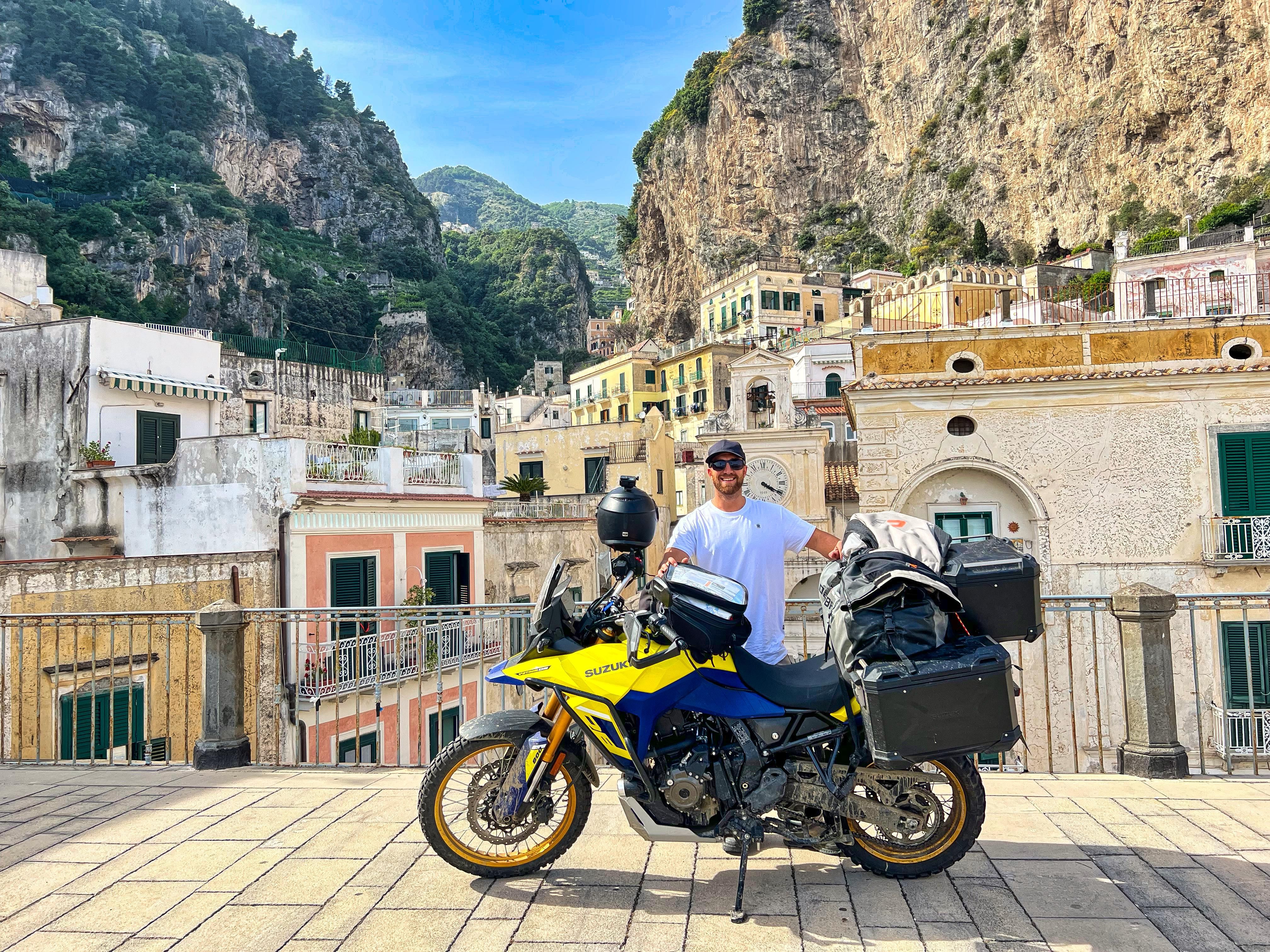 Gelb-blaues Suzuki Motorrad vor einem italienischen Bergdorf, ein Mann mit Sonnenbrille dahinter