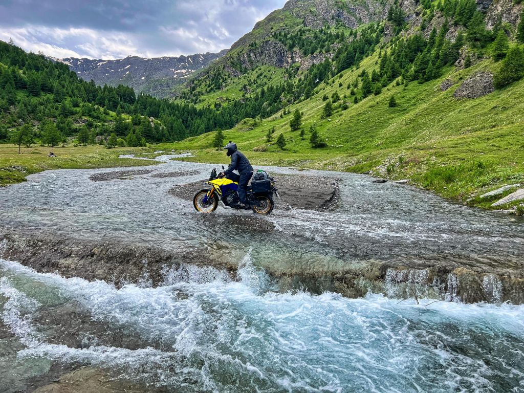 Gelb-Blaues Suzuki Motorrad bei der Durchquerung eines kleinen Flusses in einem malerischen Tal