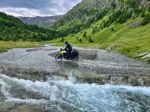 Gelb-Blaues Suzuki Motorrad bei der Durchquerung eines kleinen Flusses in einem malerischen Tal