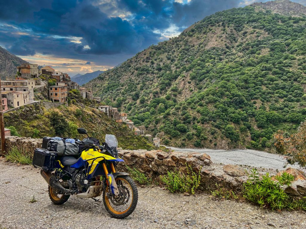 Gelb-Blaues Suzuki Motorrad vor einem italienischen Bergdorf mit Blick auf die Berge