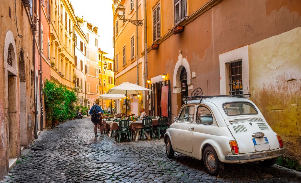 Ein malerisches italienisches Dorf, im Vordergrund ein alter Fiat 500 und dahinter ein Restaurant mit Außengastronomie