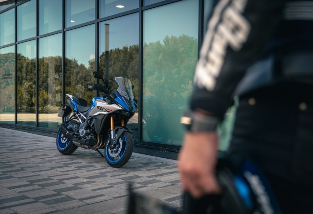 Motorradfahrer vor einem modernen Gebäude mit geparktem Suzuki-Motorrad im Hintergrund.