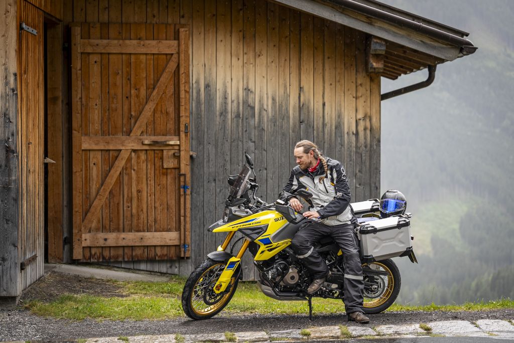 Motorradfahrer lehnt entspannt an seiner gelben Suzuki vor einer Holzhütte.