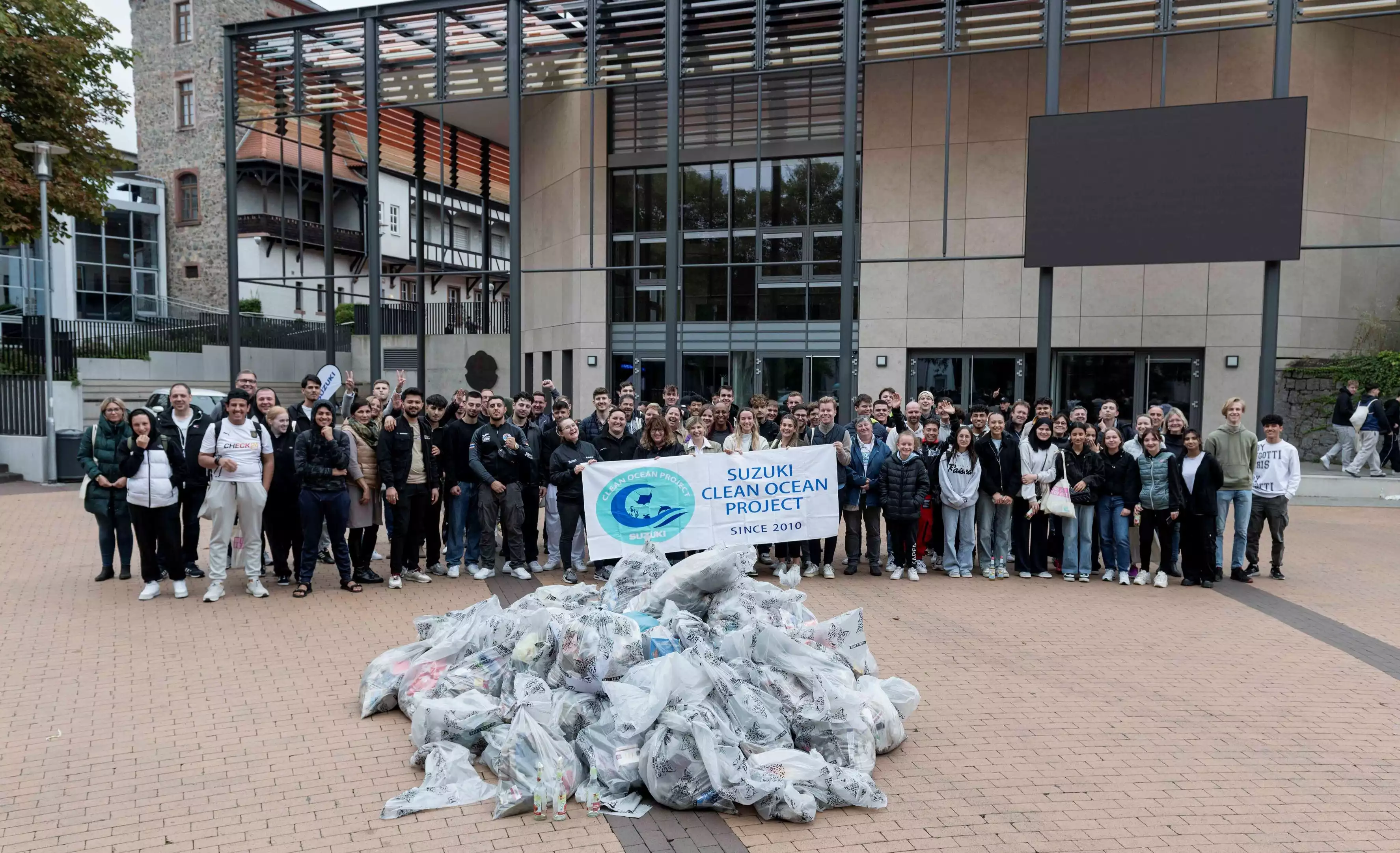 Gruppenfoto von vielen Menschen vor einem Gebäude, die im Rahmen des ‚Suzuki Clean Ocean Project‘ gesammelten Müll in Plastiksäcken präsentieren