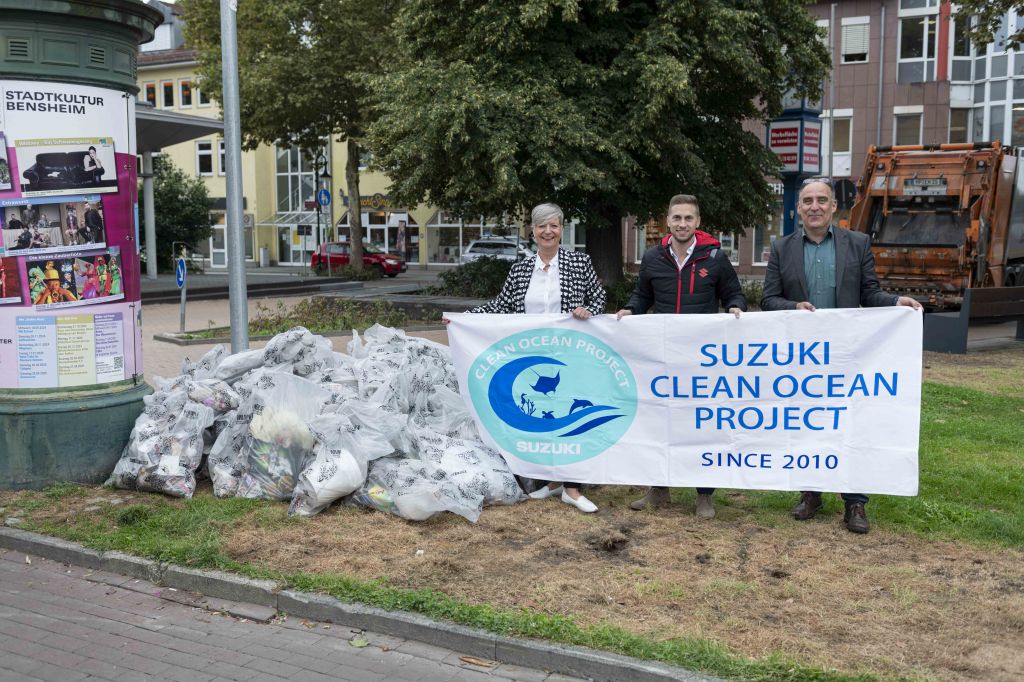 Drei Personen stehen hinter einem Banner des ‚Suzuki Clean Ocean Project‘ neben einem großen Haufen gefüllter Müllsäcke