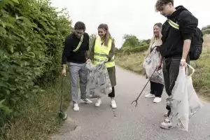 Vier Personen sammeln Müll am Wegesrand; eine Frau hält einen Müllsack, während ein Mann mit Greifzange Abfall aufnimmt