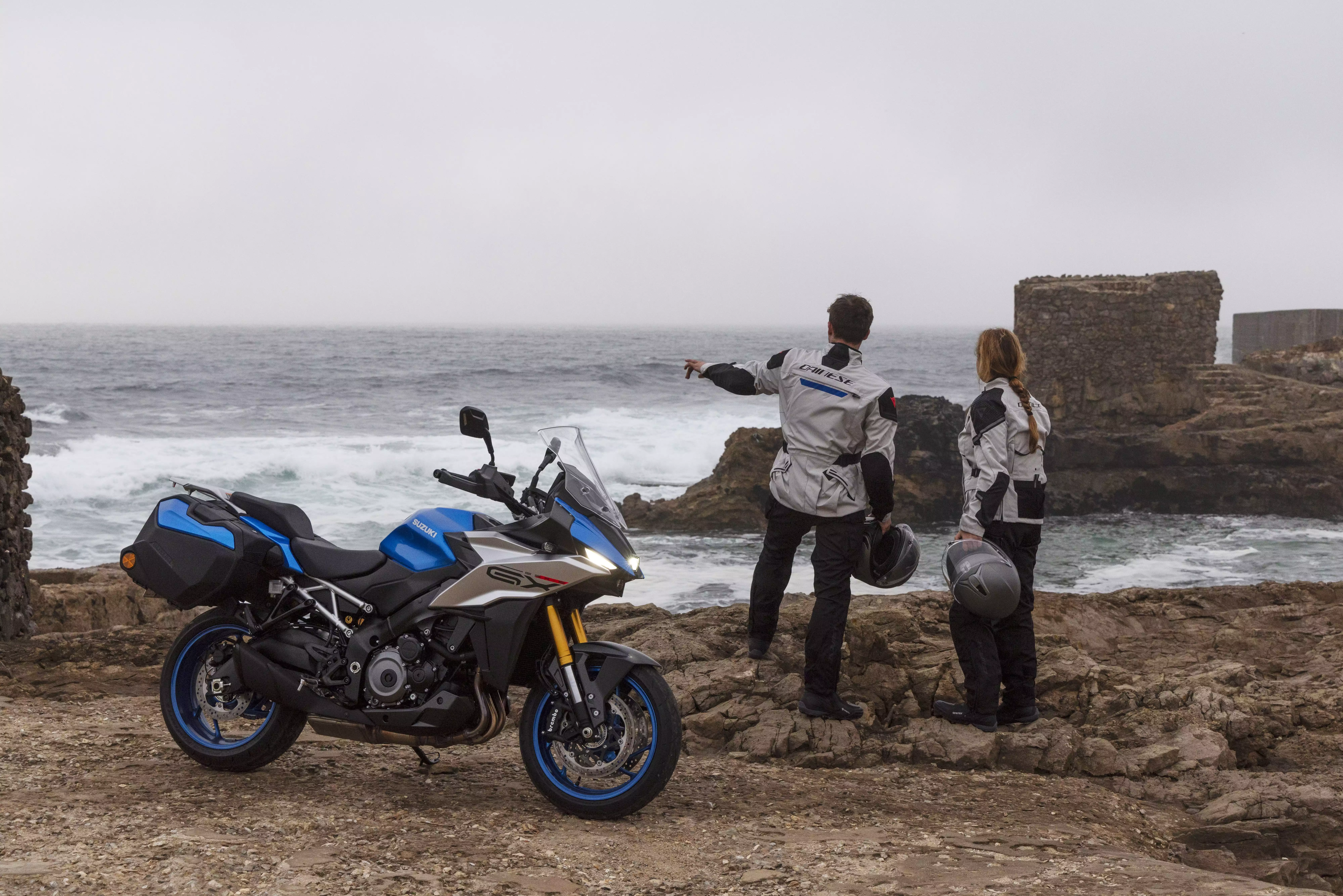 Zwei Motorradfahrende in Schutzkleidung stehen am Meer und blicken auf die Wellen; im Vordergrund steht ein Suzuki-Motorrad