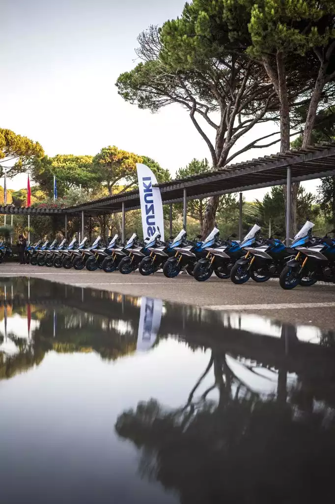 Reihe neuer Suzuki-Motorräder nebeneinander aufgestellt, mit Bäumen und Spiegelung im Wasser im Vordergrund