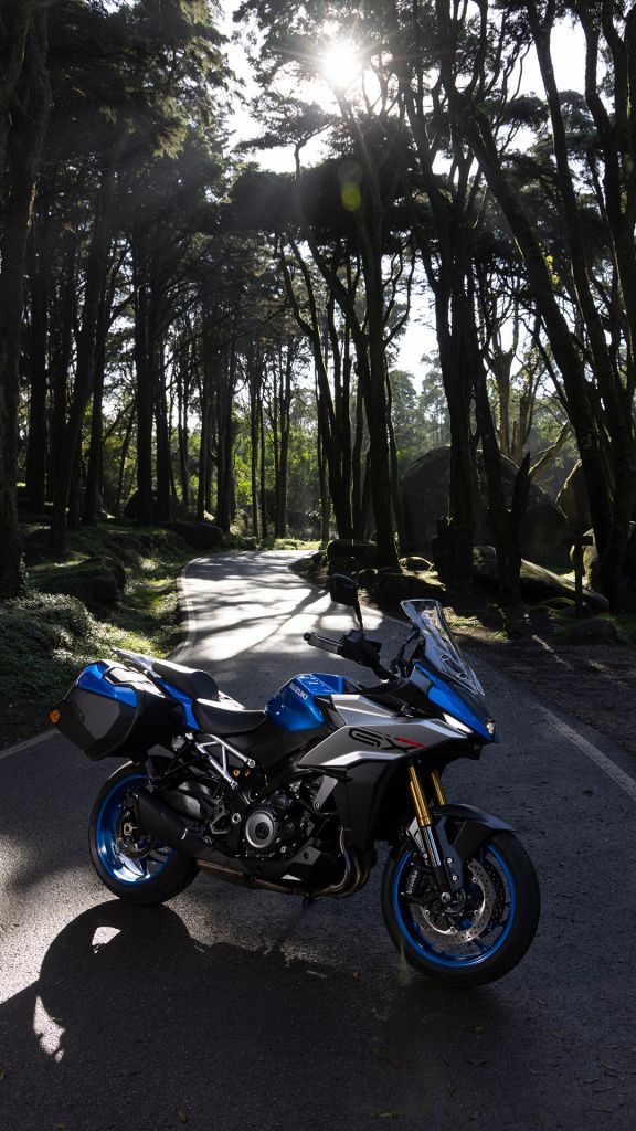 Suzuki-Motorrad steht auf einer Waldstraße, Sonnenstrahlen brechen durch die hohen Bäume im Hintergrund