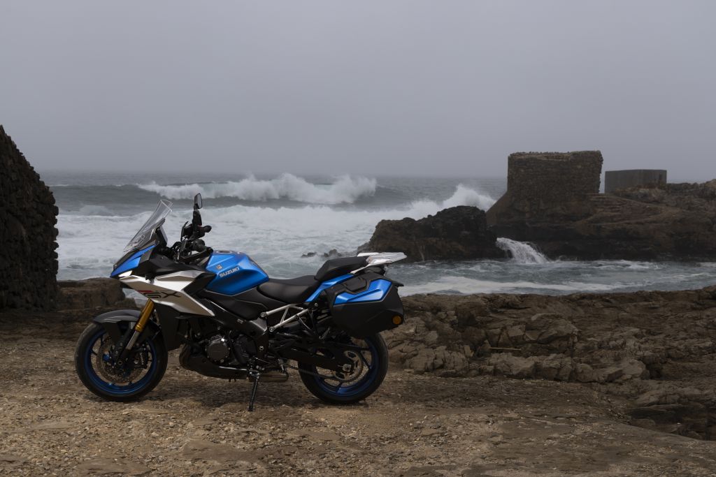 Suzuki-Motorrad steht auf felsiger Küste vor dem Meer mit Wellen und alten Steinmauern im Hintergrund